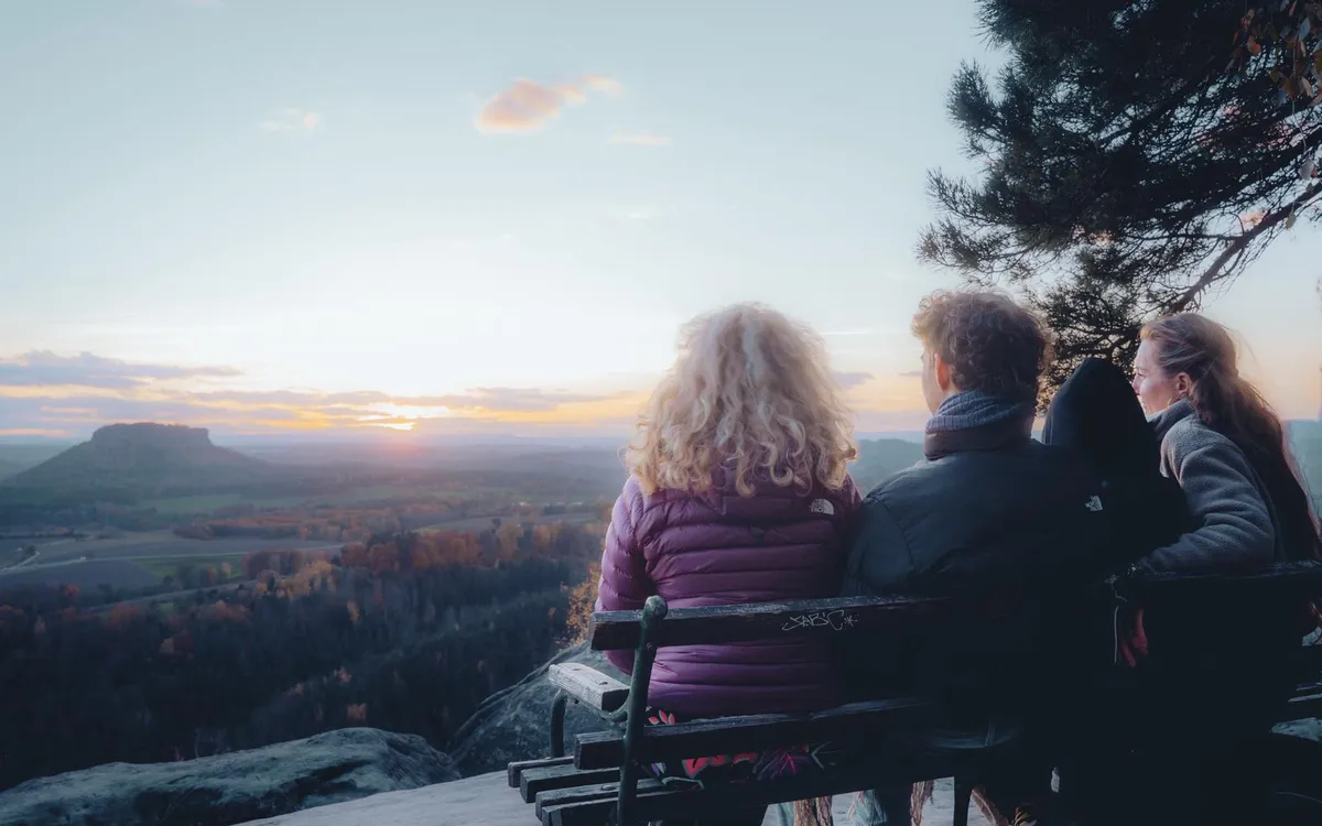 Drei Menschen sitzen auf einer Bank am Felsen und blicken in den Sonnenuntergang über dem Elbsandsteingebirge